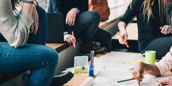 A group of young adults with pens in their hands work together on a group project and have pieces of paper spread on a table in front of them.