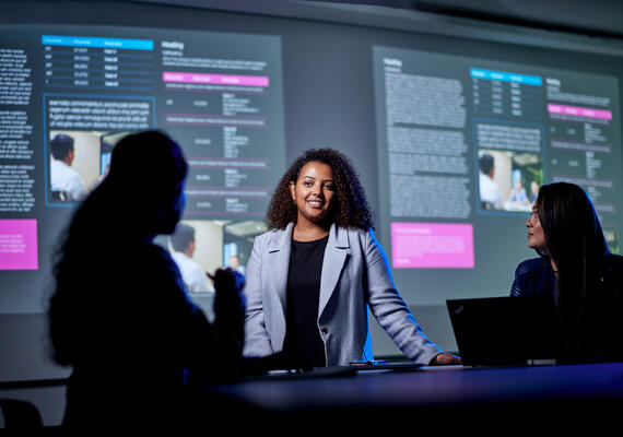 Woman presenting report to colleagues. Report information projected on screen in background.