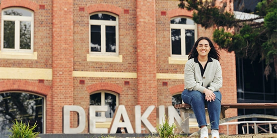 Student sits on bench outside the Deakin waterfront campus with big Deakin letters in the background.