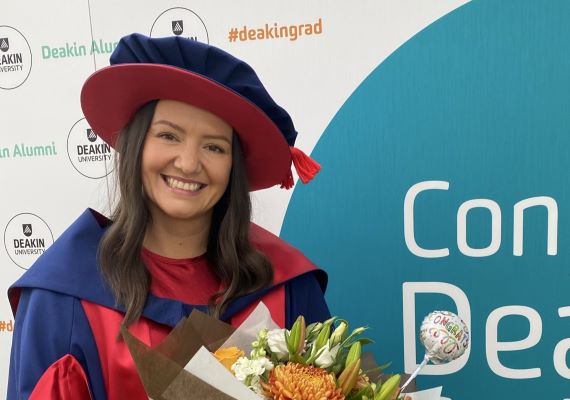 Deakin PhD student Michelle standing and smiling in graduation cap and gown whilst holding flowers.