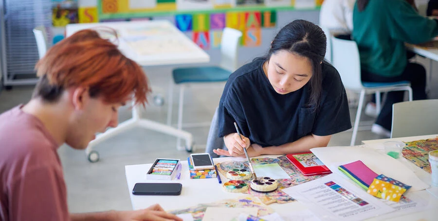 Two students sitting at a table in a classroom, using paints and crayons to practice teaching children art