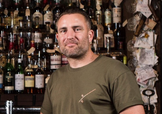 Andrew Fitzgerald, MBA graduate, co-founder and managing director of The Gospel Distillers Pty Ltd, wears green t-shirt while standing in front of a wall of whisky.