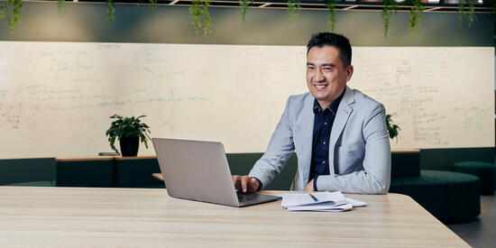 A Deakin student smiling while working on a laptop in a modern workspace, seated at a large wooden table with notebooks and a whiteboard filled with notes in the background.