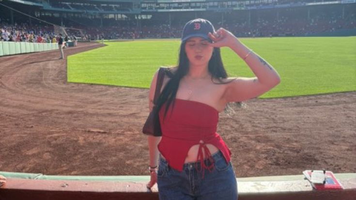 Tarnie poses in front of a baseball field. She is wearing a red top, and a blue baseball cap with a red 'B' on it.