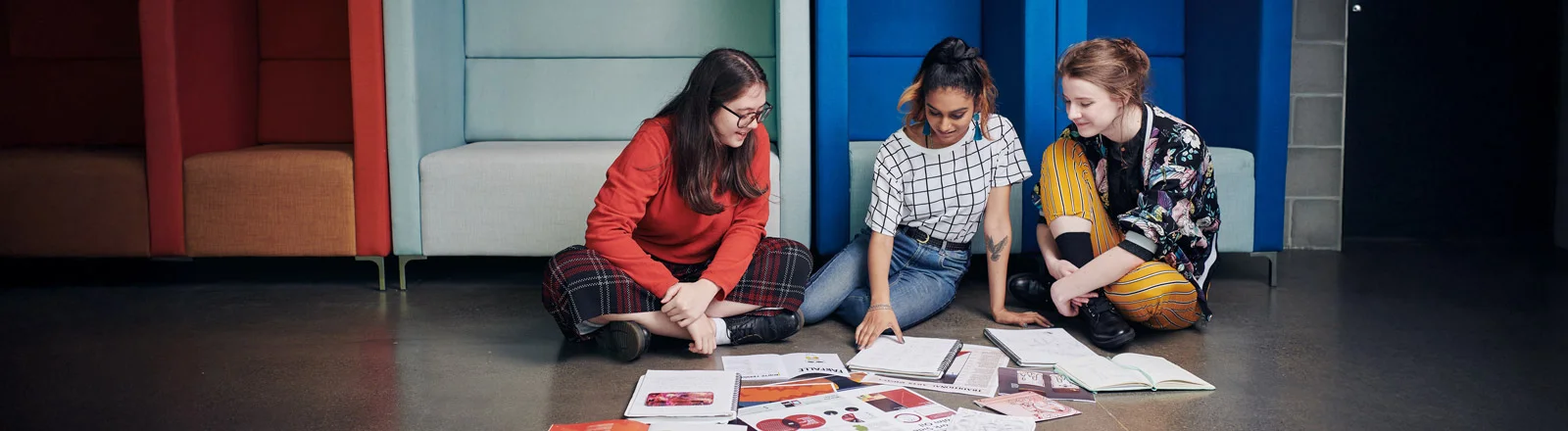 Three students studying on floor cross legged all looking at middle student's workbook smiling.