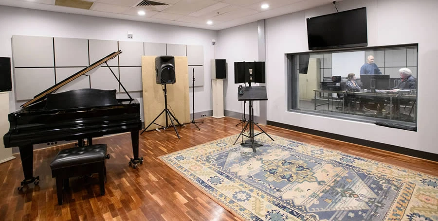 Within the sound studio, showing piano and microphones, and looking through to where people are in a control room