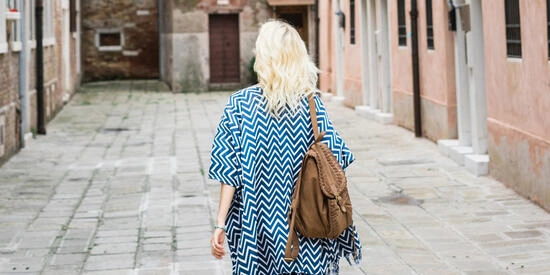 The view of a woman walking away. She has blond hair and is wearing a blue and white striped dress and backpack.