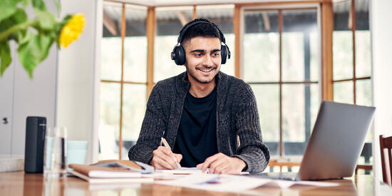 A student wearing headphones studying at home at a dining table.