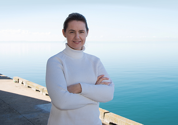 Carmel Sefian stands on a jetty with the ocean behind smiling into the camera