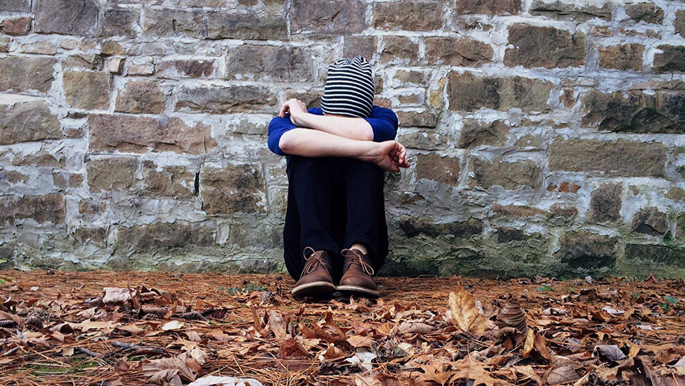 A person sitting on the ground in front of a brick wall. They have their head resting on their knees, arms crossed in front. They are surrounded by brown leaves.