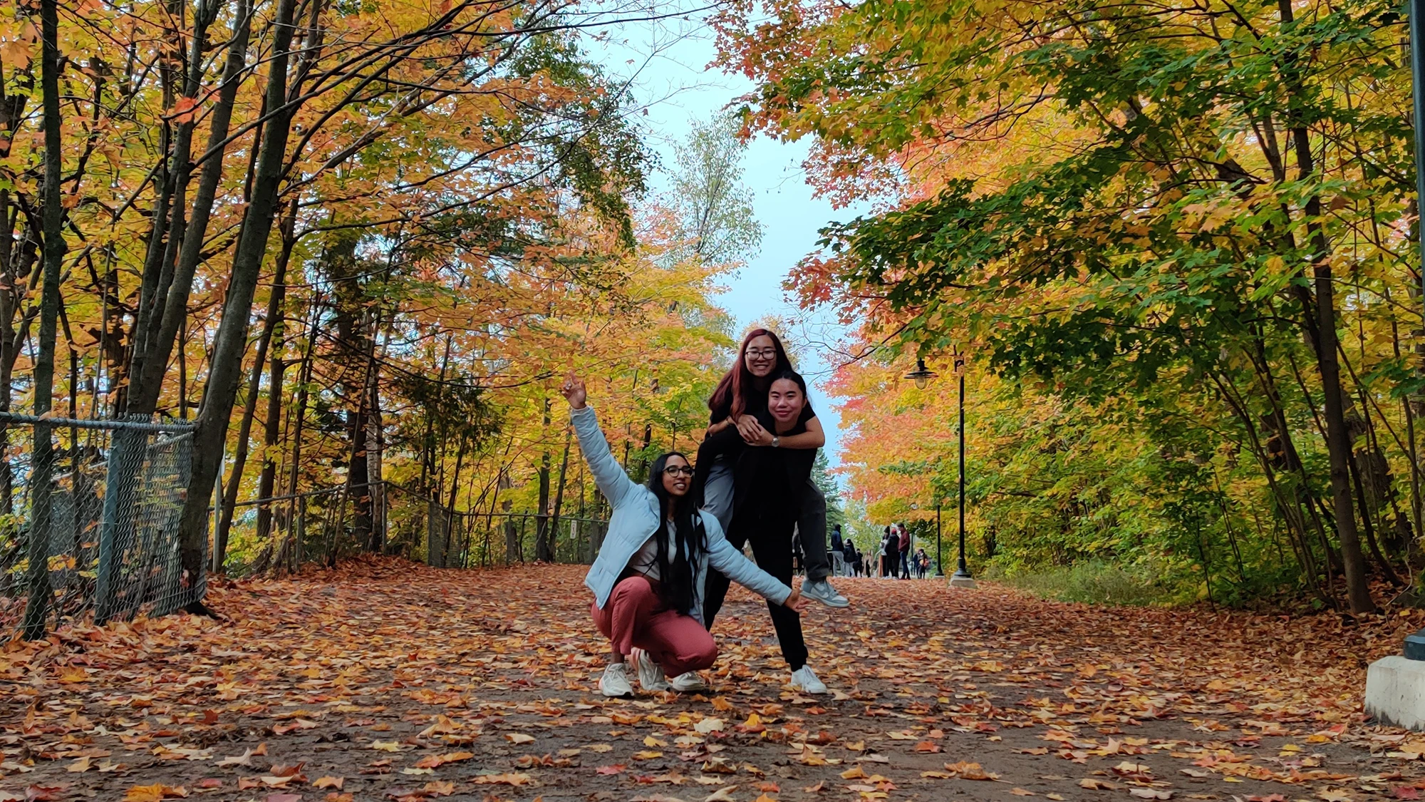 Kaviya and two friends pose in a park. The ground is covered in fallen autumn leaves, and the background is filled with trees with green, yellow, orange and red leaves.