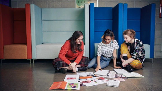 Three students studying on floor cross legged all looking at middle student's workbook smiling.