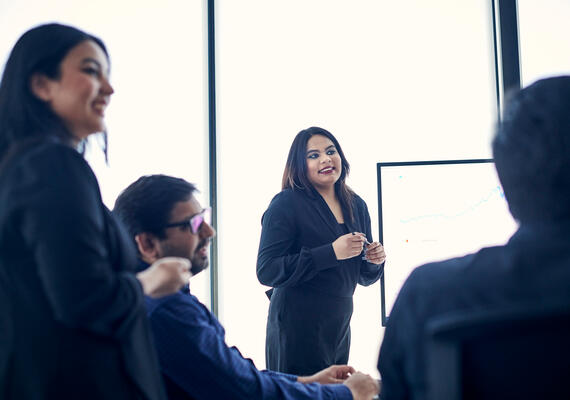 Students in a corporate meeting with one student standing next to a screen presenting.