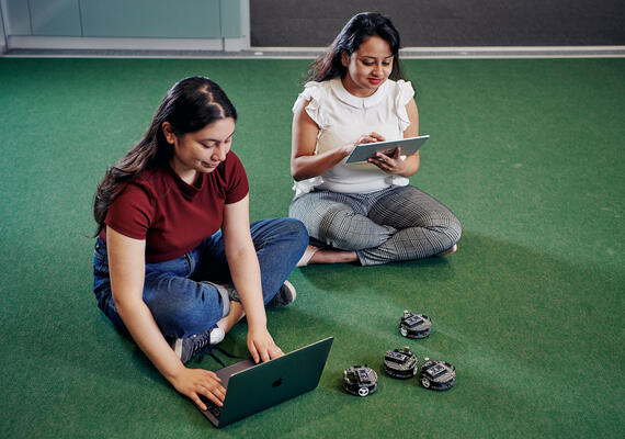 Two students control four small educational robots using a laptop and tablet computer