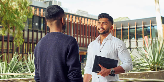 Two students stand talking. One is holding a laptop.