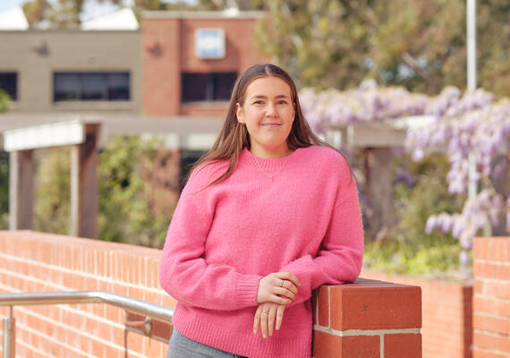 Deakin student profile image of Maya Tolliday leaning on a brick wall, smiling, on the Geelong Waterfront Campus.