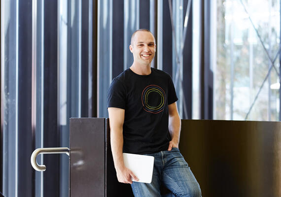 Justin Fench leans up against a wall holding a closed laptop