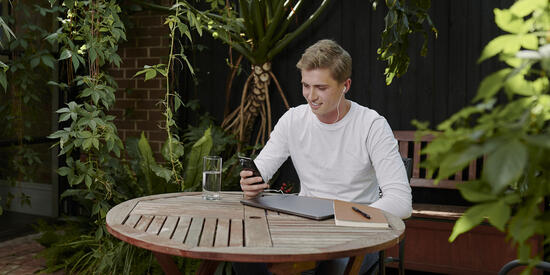 Deakin student sitting at ta table with laptop and book whilst looking at their phone.