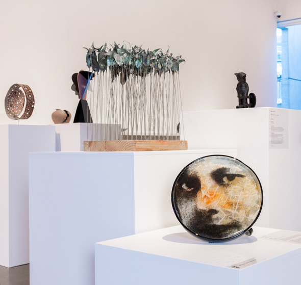 59287_590x599_contemporary_small_sculpture_award Photograph of a section of the Deakin University Contemporary Small Sculpture Award 2025 installation. In the foreground is a glasswork featuring a young face looking out at the viewer, behind is an artwork construction from metal and wood depicting an abstract forrest, in the far background are an array of other sculptures.