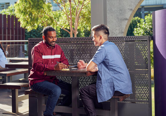 Informal interaction between two young students on campus, sitting at a table outside. 