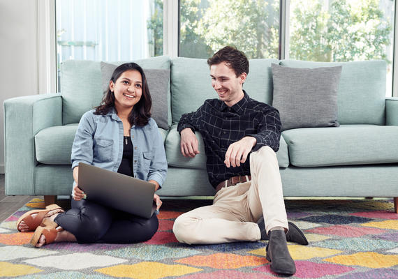 Two Deakin students sit on the floor in front of a couch, looking at a laptop.