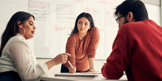 Three students in a meeting room, surrounded by papers, with notes written on the whiteboard behind them.
