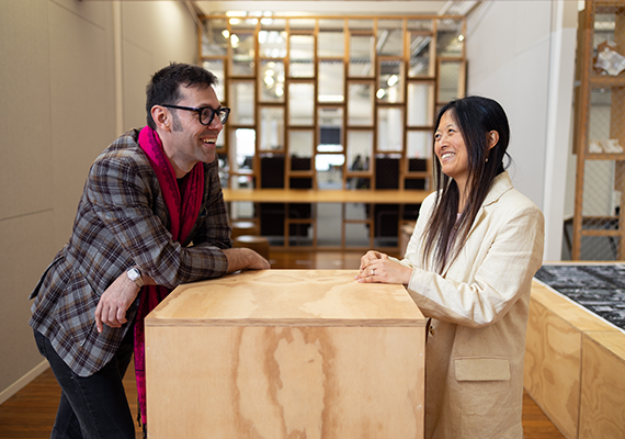 Two individuals standing across from the other, leaning against a large plywood block smiling.