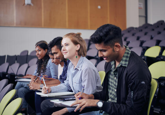 A group of students sitting in a row in a lecture theatre with study materials.