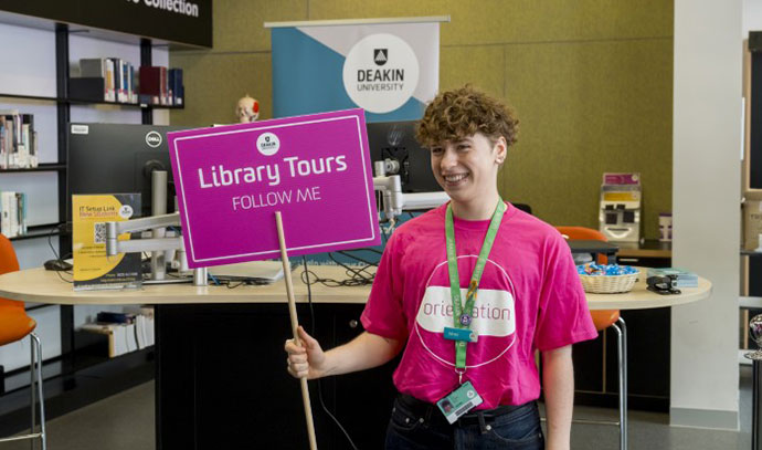 sensorytour A smiling staff member in a pink Orientation t-shirt holding a Library Tours sign