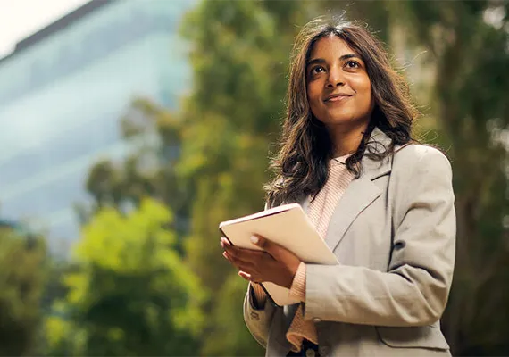 A Deakin postgraduate student standing in front of some trees, holding a notebook.