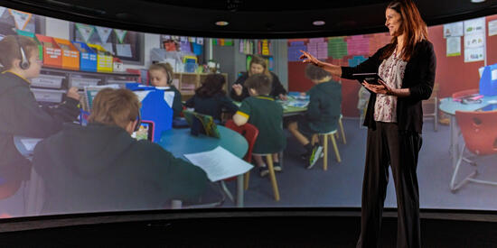 Dr Maria Nicholas Senior Lecturer in Education stands in front of screen depicting education setting.