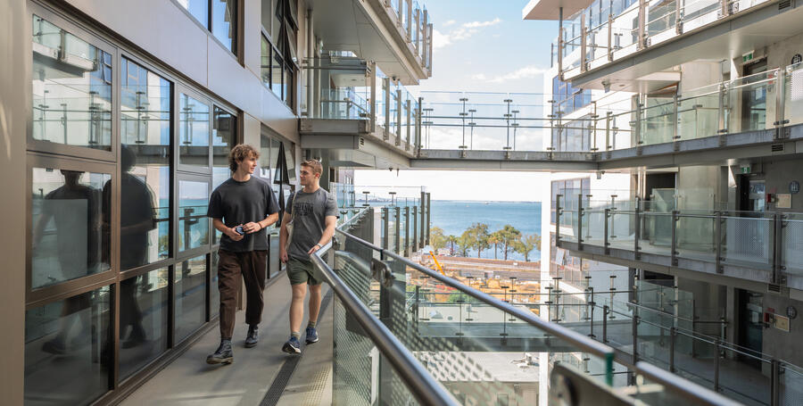 Two students walking down the path from their room at Geelong Waterfront on-campus accommodation, with the ocean in the background.