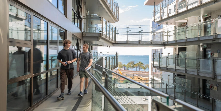 Two students walking down the path from their room at Geelong Waterfront on-campus accommodation, with the ocean in the background.