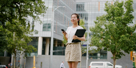 Female student standing outside with a mobile phone and a compendium under her arm.