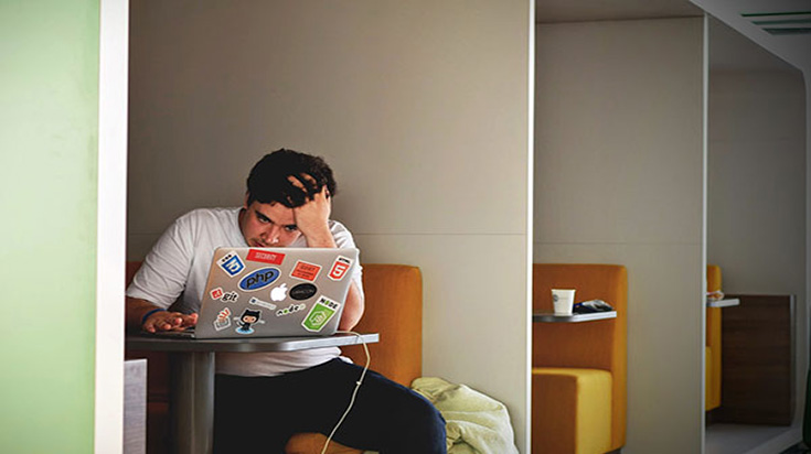 A student sitting in a library booth, looking at their laptop. They are holding their  head in their hands, looking very stressed.