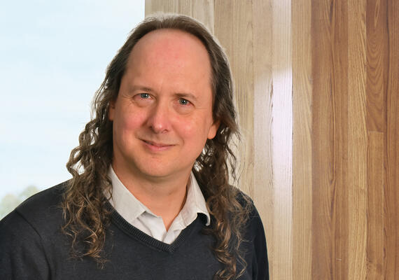 Portrait photo of male academic staff member, smiling, casually dressed, against a wood panelling background.