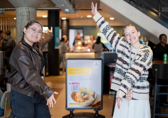 Two DUSA student council members stand in front of a sign that shows a curry dish with the title '$5 student meals' in front of a cafe area
