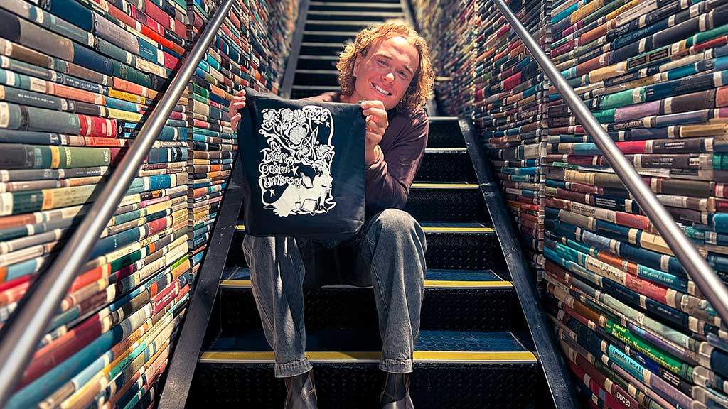 A student sitting on the internal library stairs. The walls of the stairs are made out of old library books. The student is holding a library bag showing the artwork they designed.