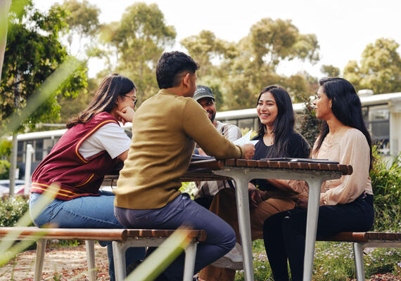 Group of students sit at an outdoor table, laughing and chatting about study.