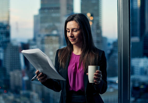 A woman stands holding papers and a mug with a city sky line behind her.