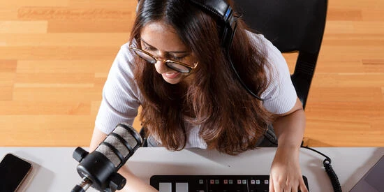 A student sitting at a desk with headphones, a microphone and computer, producing a podcast.
