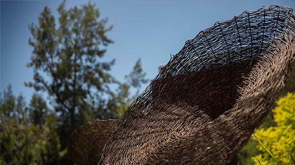 Woven basket style sculpture outside with trees and sky in background.
