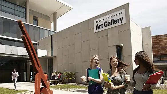 A group of three students mingle as they carry their books and walk past the Art Gallery.