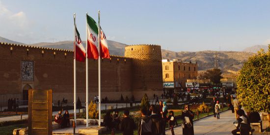 Three Iranian flags fly next to a government building, with people walking outside