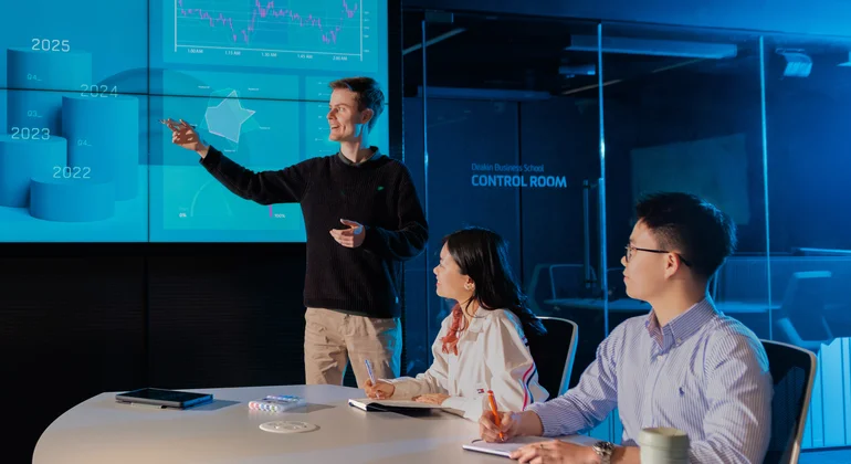 A student making a presentation in front of a large screen showing business data.
