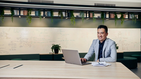 Postgraduate student working at a table inside a study area smiles away from laptop screen with reading materials by his side.