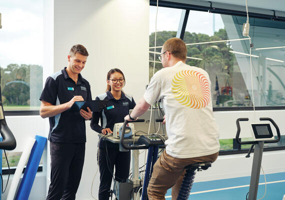 Three people inside Deakin's clinical exercise centre - one using exercise machine and two student exercise physiotherapists.