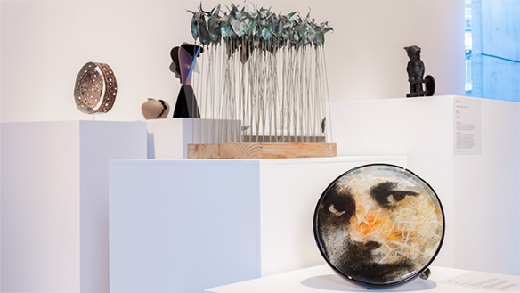 Photograph of a section of the Deakin University Contemporary Small Sculpture Award 2025 installation. In the foreground is a glasswork featuring a young face looking out at the viewer, behind is an artwork construction from metal and wood depicting an abstract forrest, in the far background are an array of other sculptures.