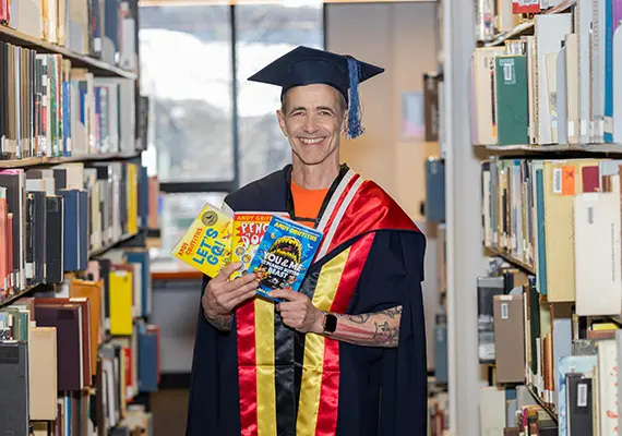 Deakin University alum and celebrated author Andy Griffiths at the September graduation ceremony at the Geelong Waterfront Campus where he was a guest speaker. He is standing in the Deakin Library, wearing his academic regalia and holding some of his best-selling books.