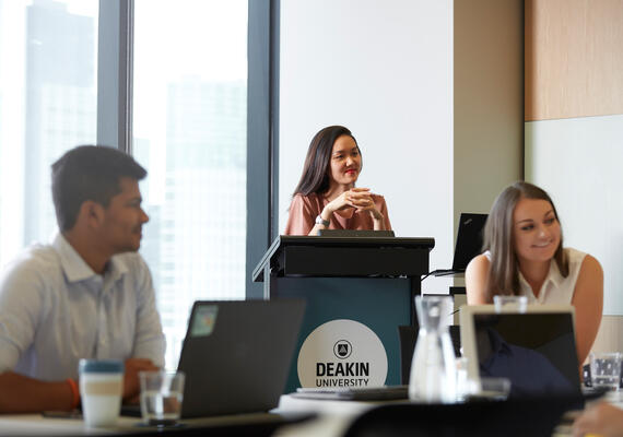 Group of Deakin professionals in a meeting room. One stands at a lectern, two sit at a table.